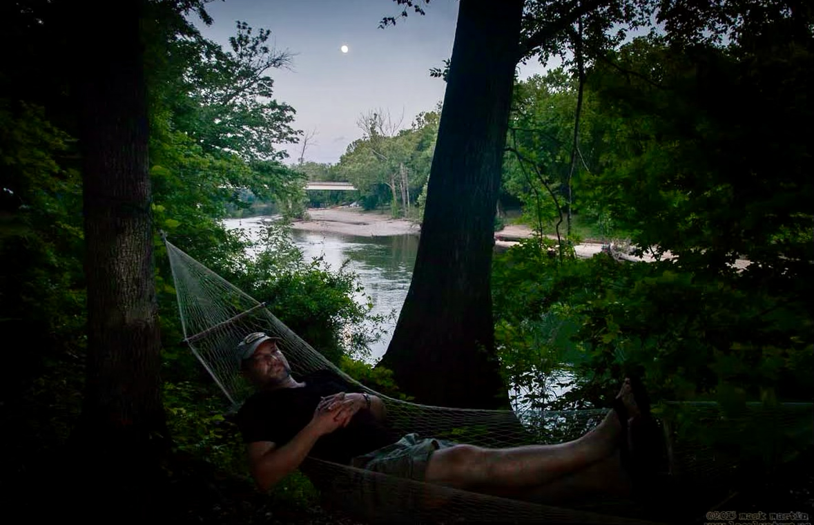 Camper resting in a hammock beside the Buffalo River as the moon rises over Bell Bridge