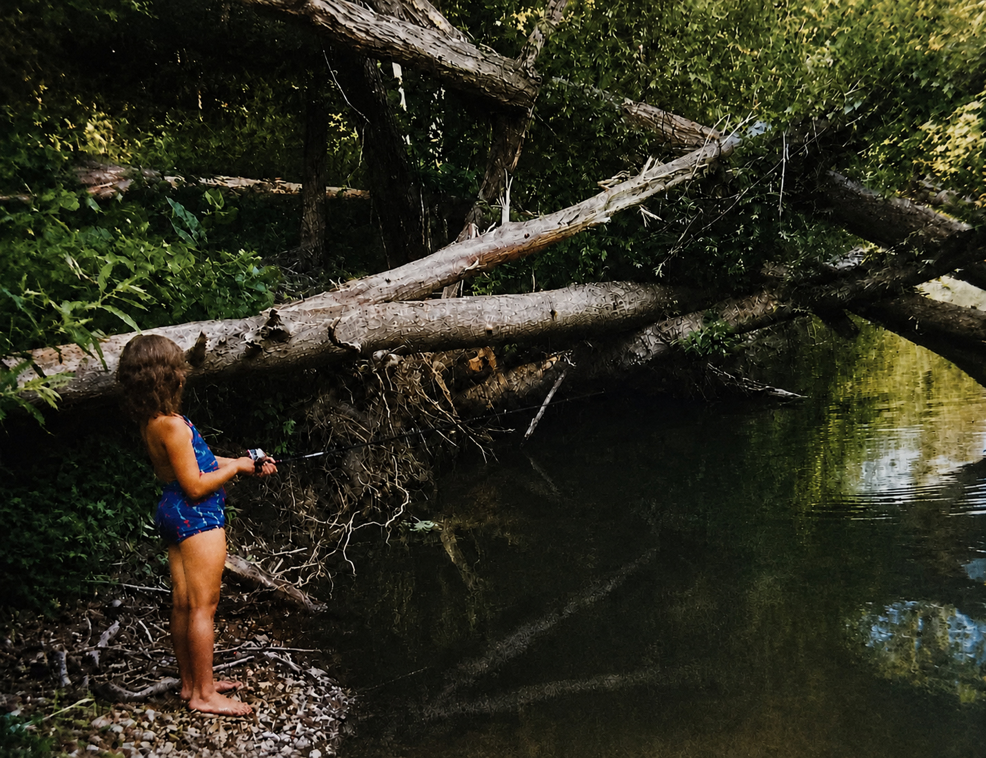 A young girl in a swimsuit fishing from the gravel bank beside a fallen tree on the Buffalo River in the 1990s